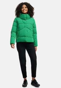 Giacca a vento verde con colletto alto, caratterizzata da sezioni trapuntate e bottoni a pressione. Pantaloni neri slim-fit e sneakers scure completano l'outfit.
