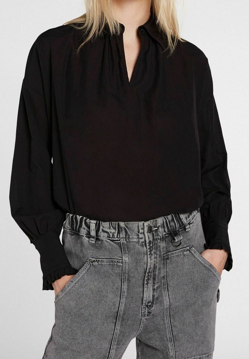 Femme portant un chemisier noir à manches longues avec de petits volants au niveau des poignets et un jean gris clair taille haute, les mains dans les poches.