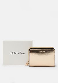 Portefeuille métallique doré de Calvin Klein avec fermeture éclair, texture lisse et accent avec branding. Présenté dans une boîte blanche simple.