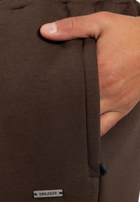 Braune Baumwoll-Jogginghosen mit einer Seitentasche. Sie haben eine glatte Textur und ein kleines Metallschild mit der Aufschrift "SMILODOX" am Saum.