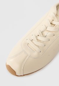 Baskets en cuir crème avec un bout arrondi, des lacets plats et des détails de couture subtils. Semelle extérieure en caoutchouc brun au design minimaliste.