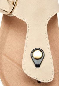 Sandalia de ante beige que presenta un tira de dedo triangular, herrajes negros y un acento circular dorado. Textura suave con detalles de costura finos.