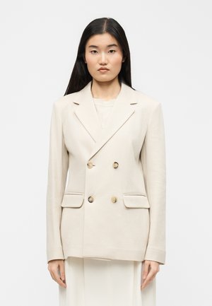 Žena s dlhými čiernymi vlasmi, oblečená v svetlobéžovom dvojradovom saku s dvoma prednými vreckami, stojaca pred jednoduchým bielym pozadím.