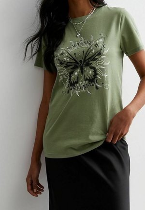 Nainen pukeutunut oliivinvihreään t-paitaan, jossa on mustavalkoinen perhonsilta, ja mustaan hameeseen, asusteena hopeinen sydänkaulakoru.