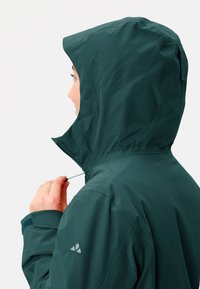 Giacca impermeabile verde scuro con cappuccio, coulisse regolabile e polsini elastici. Presenta un logo sulla parte superiore della manica sinistra.