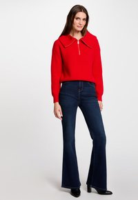 Rode gebreide trui met een grote kraag en ritssluiting, gecombineerd met donkerblauwe flared jeans en zwarte haklaarzen. Eenvoudig en modern ontwerp.