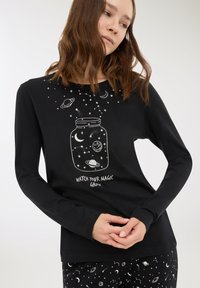 Donna che indossa una maglietta a maniche lunghe nera e pantaloni con stampe spaziali bianche e il testo "Guarda crescere la tua magia" sulla maglietta.