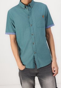 Kortärmad skjorta med knappar i teal-färgat tyg, med en bröstficka, kontrasterande blå manschetter och bruna knappar. Strukturerat utförande.