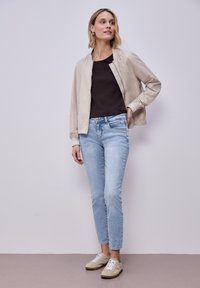 Kobieta w beżowej kurtce bomber, czarnym topie, jasnoniebieskich jeansach i beżowych trampkach, stojąca na tle gładkiej jasnej ściany.