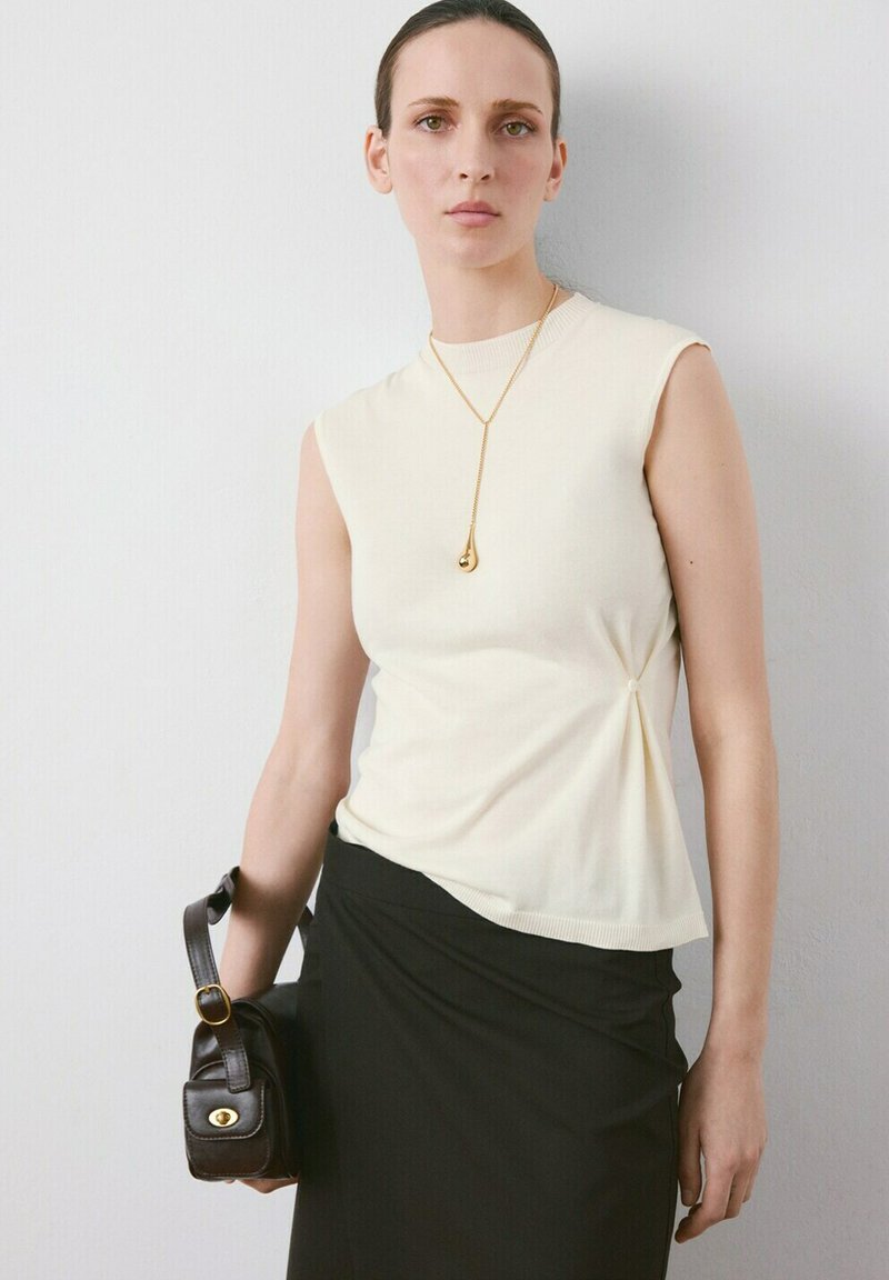 Žena u kremasti majici bez rukava s detaljem sa strane, crnoj suknji, zlatnoj ogrlici s privjeskom i drži malu tamnosmeđu torbicu.