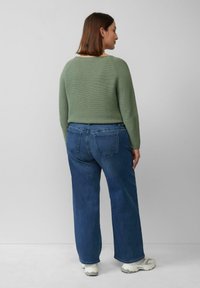 Groene, getextureerde trui met een ronde halslijn, gecombineerd met hoge blauwe spijkerbroeken. Outfit compleet met lichte sneakers.