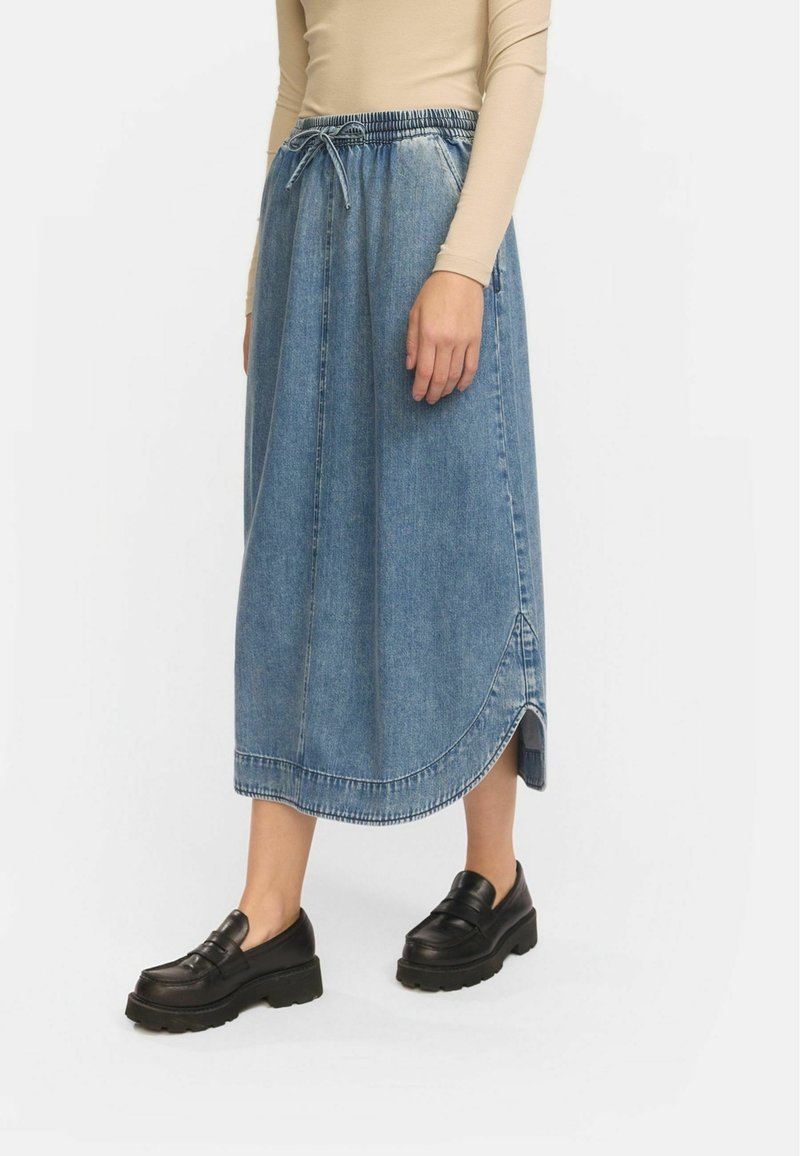 Denim maksi krilo, svetlo modro, z elastičnim pasom in vrvico. Ima ukrivljen rob in stranske razpoke. Nosi se z črnimi platform čevlji.