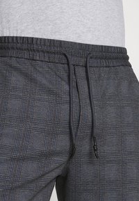 Tummansininen ja harmaa ruudullinen verkkarit, joissa on joustava vyötärö, säädettävä kiristysnauha ja teksturoidulla kuviolla varustettu kangas.