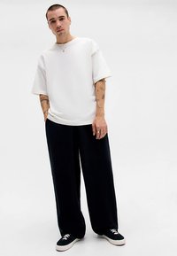 Moški s kratkimi lasmi, oblečen v belo preveliko majico, široke črne hlače in črne superge, stoji z rokami v žepih na enobarvni beli podlagi.