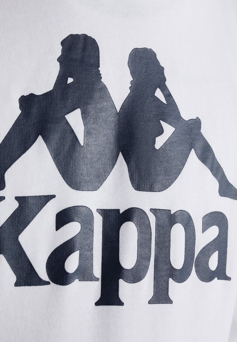 Camiseta blanca con un gran logo negro de "Kappa" y siluetas de dos figuras sentadas, con textura suave y diseño de cuello redondeado.