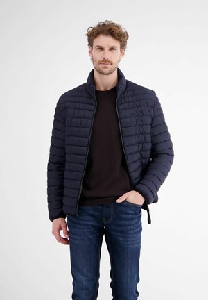 Man met lichtbruin krullend haar die een zwarte donzen jas, zwart shirt en blauwe spijkerbroek draagt, staand met één hand in zijn zak.