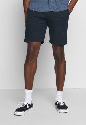 Shorts blu navy in cotone con taglio dritto, orlo arrotolato, indossati con sneakers nere e calzini bianchi. Design semplice, senza fantasie.