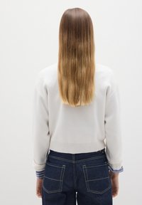 Weißer Pullover mit geripptem Saum, getragen über einem blau-weiß gestreiften Hemd. Kombiniert mit dunkelblauen Jeans mit doppelten Gesäßtaschen.