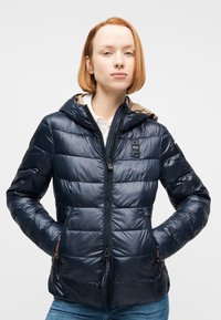 Νέος με κοντά κόκκινα μαλλιά, φορόντας μια σκούρα μπλε φουσκωτή μπουφάν με κουκούλα και τα χέρια στις τσέπες, στέκεται μπροστά σε απλό φόντο.