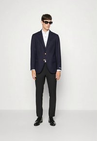 Άνδρας που φοράει μαύρα γυαλιά ηλίου, ναυτικό σακάκι με χρυσά κουμπιά, λευκό πουκάμισο, μαύρο παντελόνι και γυαλιστερά μαύρα παπούτσια, στέκεται σε απλό φόντο.