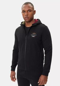 Felpa nera con zip realizzata in tessuto morbido, dotata di tasca a marsupio, polsini a coste e colori contrastanti sulla fodera del cappuccio: bordeaux e verde.