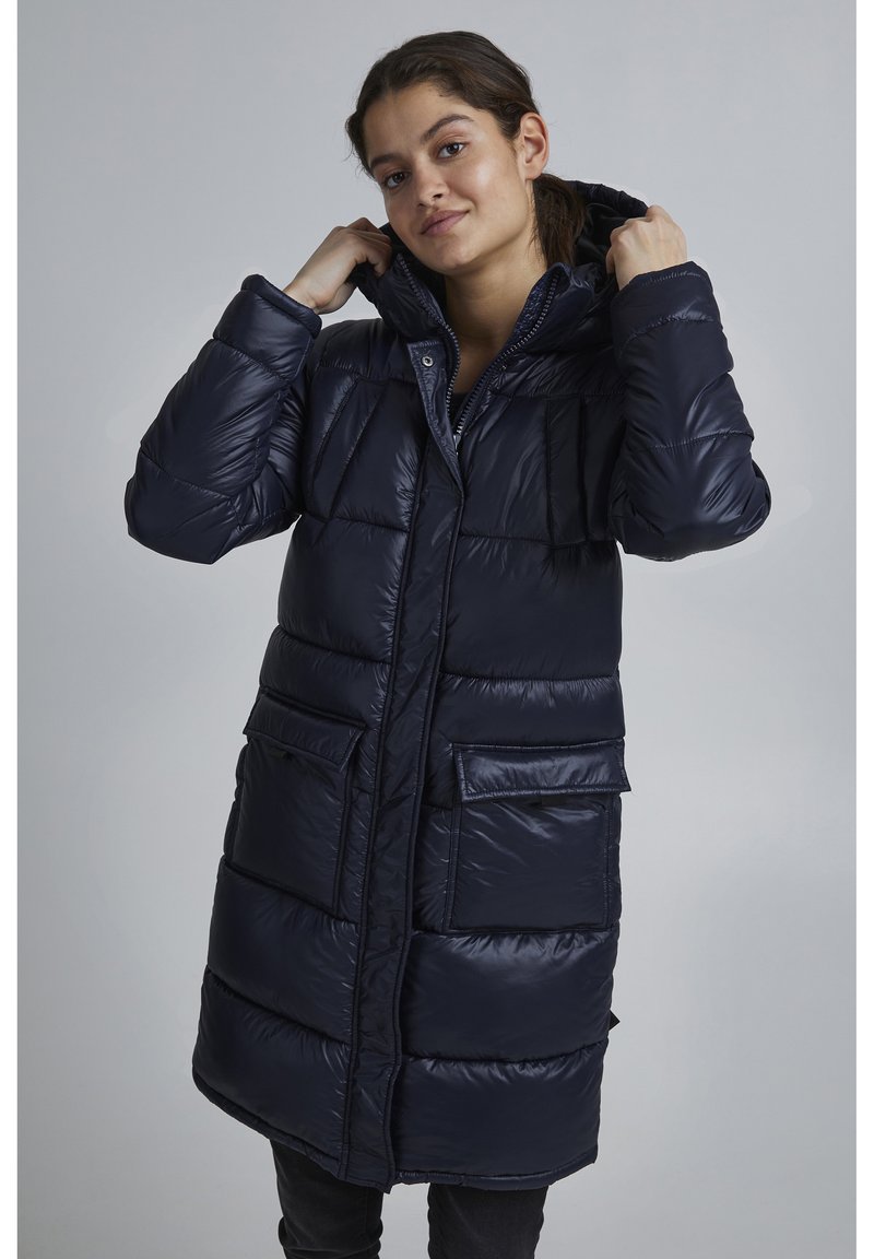 Manteau matelassé long, bleu marine, avec capuche. Doté d'une fermeture éclair à l'avant, de grandes poches plaquées et d'un design matelassé avec une texture lisse et brillante.