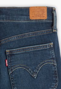 Jean en denim bleu foncé avec une étiquette en cuir à la taille, présentant des coutures contrastées et un design distinctif de la poche arrière.