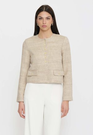 Donna con lunghi capelli scuri che indossa una giacca beige texturizzata con bottoni e pantaloni bianchi, in piedi davanti a uno sfondo chiaro e uniforme.