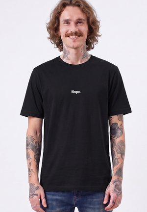 Jeune homme avec des tatouages et un piercing au septum souriant, portant un t-shirt noir avec le mot "Nope." imprimé sur la poitrine.