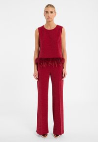 Rode twee-delige outfit met een mouwloze top met glinsterend effect en verenafwerking, gecombineerd met aansluitende uitlopende broek gemaakt van hetzelfde materiaal.