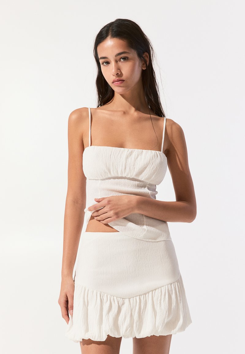 Ensemble blanc en deux pièces comprenant un haut court texturé avec des bretelles fines et une mini-jupe à volants, mettant en valeur un tissu doux et transparent.