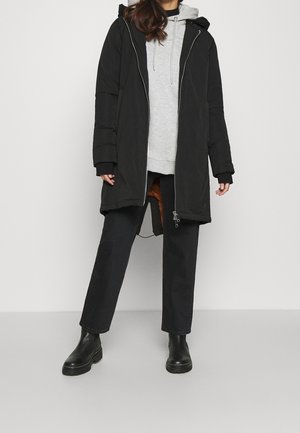 Personne portant un parka noir à capuche par-dessus un sweat à capuche gris, un pantalon large noir et des bottines noires, sur un fond clair uni.