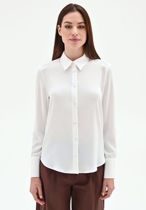 Femme portant un chemisier blanc à manches longues boutonné et un pantalon marron, debout devant un fond uni et clair.