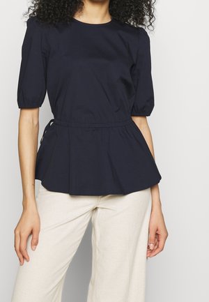 Vrouw draagt een marineblauwe blouse met korte mouwen en geplooide taille, gecombineerd met crèmekleurige broek, staand tegen een effen lichte achtergrond.