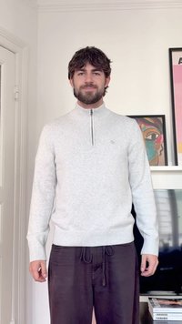 Hombre con cabello oscuro que lleva un suéter gris claro con cremallera hasta el pecho y pantalones negros, de pie en interiores cerca de una obra de arte abstracta enmarcada y un televisor.