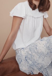 Vit bomullstopp med volangärmar och smockad halsringning, kombinerad med en ljusblå mönstrad kjol med blommönster och vågiga design.