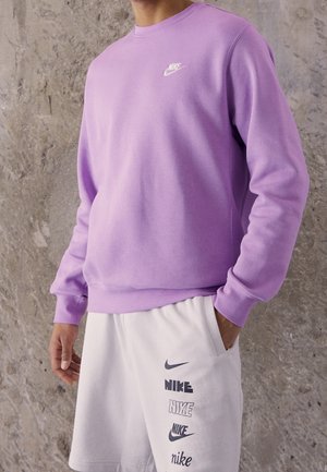 Mężczyzna w lawendowej bluzie Nike i białych szortach z wieloma czarnymi logo Nike, stojący na tle szarej, teksturowanej ściany.