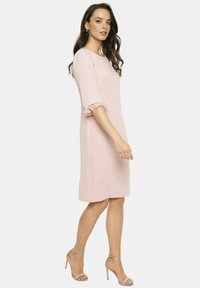 Jasnoróżowa sukienka z trzema ćwiartkami falbaniastego rękawa, do kolan, wykonana z gładkiego materiału. Modelka ma na sobie srebrne sandały na obcasie.