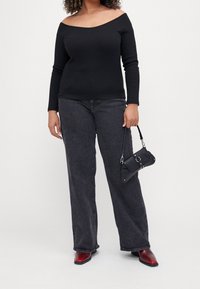 Nainen, jolla on yllä musta olkapäiltä avonainen pitkähihaisuus toppi, tummanharmaat leveälahkeiset farkut, punaiset kärjet saappaat ja hän pitää pientä mustaa käsilaukkua kahvasta.