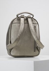 Metaliczny srebrny plecak o teksturowanej powierzchni, z podwójnymi paskami, górnym uchwytem i zapinaną na suwak kieszenią z boku. Minimalistyczny design.