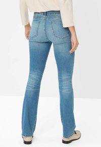 Vista posteriore di una persona che indossa jeans bootcut azzurri, una maglietta bianca e mocassini beige su uno sfondo bianco uniforme.