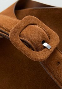 Hnědý semišový popruh na kabelku se čtvercovou přezkou. Texturovaný materiál, hladká stříbrná kovová část, minimalistický design a detaily prošití.
