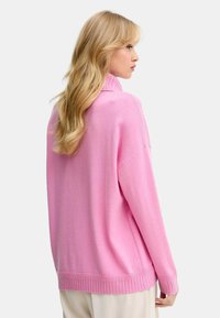 Maglione rosa lavorato a maglia con colletto alto, spalle a caduta e orlo a costine. Texture morbida, vestibilità rilassata. Nessun motivo o decorazione visibile.