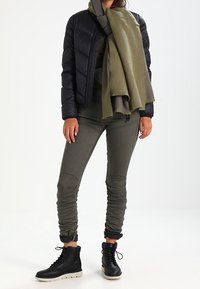 Svart pufferjacka, olivgrön scarf, mörkgröna tajta byxor med rynkningar. Svarta ankelstövlar med vita sulor. Släta texter, avslappnad design.