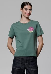 T-shirt en coton vert avec un col rond et des manches courtes, présentant un design de fleur de lotus rose sur le côté gauche de la poitrine.