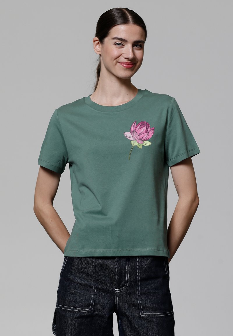 T-shirt en coton vert avec un col rond et des manches courtes, présentant un design de fleur de lotus rose sur le côté gauche de la poitrine.