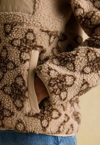 Person’s hand in pocket of beige and brown patterned fuzzy jacket, with visible blue denim jeans at waist.