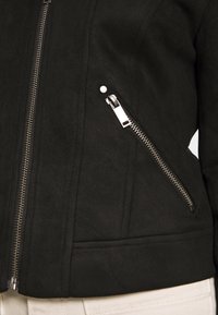 Giacca nera in pelle sintetica con un design aderente, dotata di una cerniera argentata e di una tasca laterale con chiusura lampo. Texture morbida, finitura liscia.