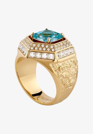 Złoty pierścionek z dużym, kwadratowym, niebieskim kamieniem szlachetnym, otoczonym małymi, przezroczystymi kamieniami, oraz z wygrawerowaną twarzą lwa na obrączce.