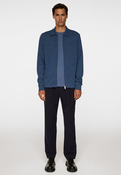 Giacca blu con zip e colletto, indossata sopra una maglietta blu, abbinata a pantaloni scuri e scarpe nere. Tessuto liscio e design minimale.
